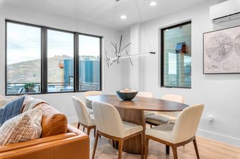 A dining room with a wooden table and white chairs, a leather couch, and windows overlooking the mountains. The room features a modern chandelier with linear lights and a framed map of the world on the wall.