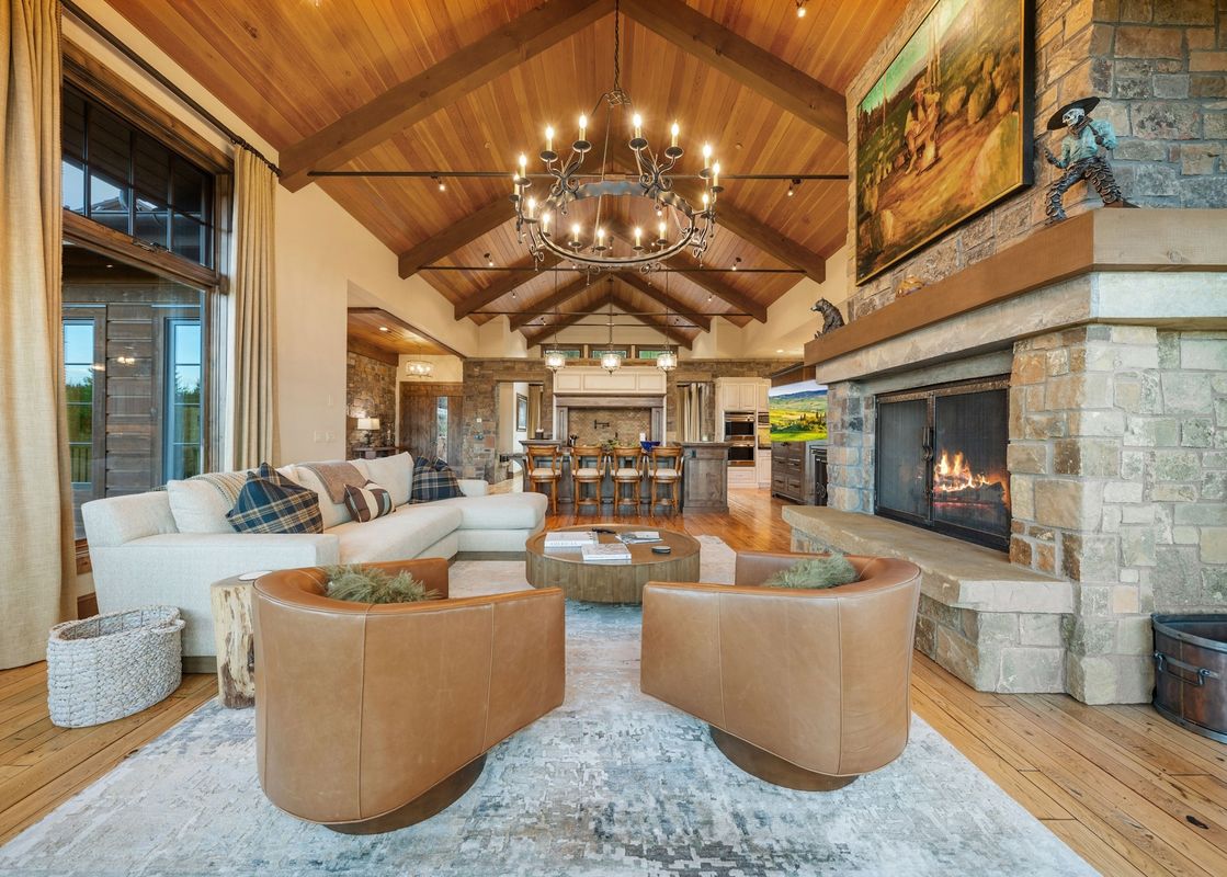 A luxurious and spacious living room with high wooden ceilings, a large stone fireplace, and elegant furnishings, including a white sectional and two brown leather armchairs.