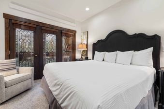 A king-sized bed with a dark wooden headboard and white bedding is positioned between a bedside table with a lamp and double doors with leaded glass panels, revealing a mountain view.