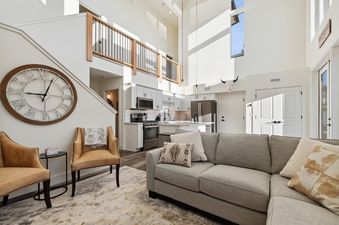 A wide-angle view of the bright and spacious living room in Quarry B23, featuring a comfortable gray sectional, two tan armchairs, a large rustic wall clock, and an open-concept kitchen in the background.