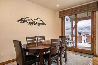 A cozy dining area featuring a wooden oval table with six dark chairs, mountain and bear-themed wall decor, and a glass sliding door leading to a snowy outdoor patio.