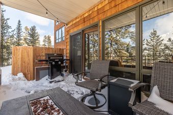 The outdoor patio of Snowflake at Skycrest features a BBQ grill, seating, and a fire pit, all surrounded by snow with a view of pine trees.