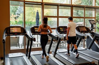 A man and woman walk on treadmills in a gym with large windows overlooking a lush green landscape.