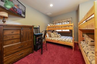 A cozy bedroom in Baldy Ridge Chalet features bunk beds with bear-themed bedding, a wooden dresser with a lamp, and an arcade machine on a red carpet.