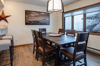The dining area at Ranch at Steamboat RA203 with a dark wood dining table, matching chairs, a breakfast bar, and a window overlooking a snowy landscape.