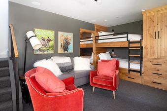 A cozy living area in Moonlight Basin 27 Lake Cabin features a red velvet armchair, a couch, and a bunk bed, with artwork featuring moose and bears on the walls. The room has a warm and inviting atmosphere.