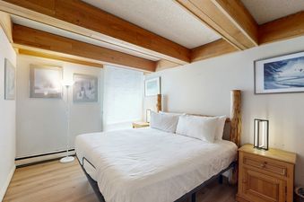 A king-size bed with white linens and a wooden headboard is centered in the bedroom of Vail International 003, flanked by a modern bedside lamp and a floor lamp.