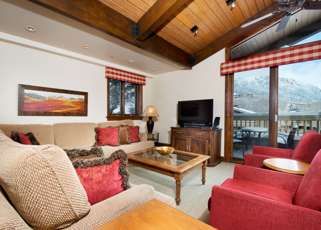 The living area of The Gant Aspen 4 Bedroom Premier features a comfortable L-shaped sofa and red armchairs, with a large glass door opening to a balcony overlooking snow-covered mountains.