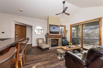 The living area within Aspen Trail Junction is warm and inviting, featuring a cozy fireplace, comfortable seating, and a large sliding glass door that offers views of the snowy outdoors.