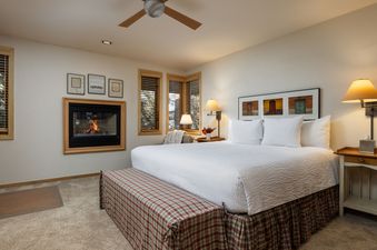 A cozy king bedroom in Tamarack 01 with a fireplace, comfortable bedding, a plaid bench, and natural light from wooden-blinded windows.