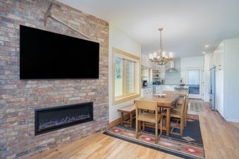 An open concept living area featuring a brick accent wall with a TV and fireplace, a wooden dining table, and a modern kitchen.