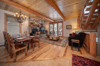 The interior of the 500 North Lupine features a dining area with a wooden table set for six, a living space with a brown sofa and a large stone fireplace, and a natural wood ceiling with exposed beams, illuminated by a chandelier and track lighting.