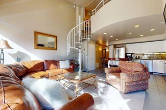 A bright and spacious living area with a brown leather sectional, glass coffee table, and a white spiral staircase leading upstairs, adjacent to a modern kitchen.