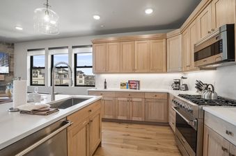 A modern kitchen with light wood cabinets, white countertops, and stainless steel appliances is shown, featuring a sink on the island and large windows with an outdoor view.