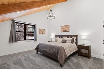 A cozy bedroom at Alpine Glow Ski Haus with a large bed, two nightstands with lamps, a window with a snowy view, and a vaulted ceiling with exposed wooden beams and a chandelier.