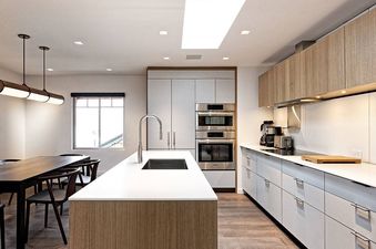 A modern kitchen and dining area with light wood cabinetry, white countertops, stainless steel appliances, a long dining table, and large window.