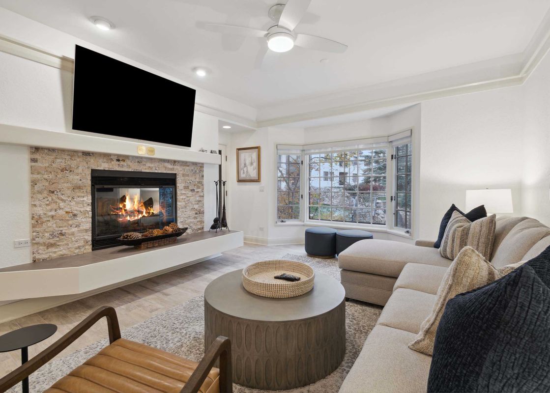 Cozy living room at The Charter C250 featuring a stone-accented fireplace, wall-mounted TV, sectional sofa, and bright bay windows.