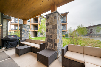 A covered outdoor patio features wicker sofas with light cushions, a matching coffee table, and a black Weber grill, all set against a backdrop of green grass, trees, and apartment buildings under an overcast sky.