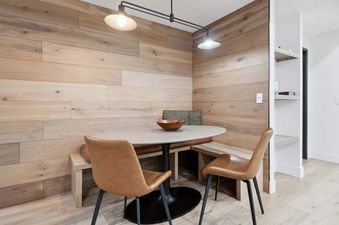A cozy dining nook featuring an oval table, leather chairs, and built-in wooden bench seating with olive-green cushions, all set against light-toned wooden panel walls under modern track lighting.