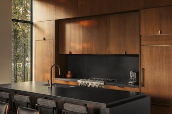 Modern kitchen at the Four Bedroom Private Home Moonlight Basin with warm wood cabinets and a black countertop island looking out a forest window.