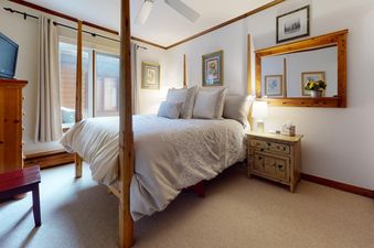 The bedroom in All Seasons 3 Bedroom Platinum Unit B4 features a large four-poster bed with white bedding, a dresser with a television, and a bedside table with a lamp, all illuminated by natural light from a large window.