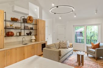 A modern and bright open-plan living and kitchen area at Depot Place, featuring light wood cabinetry, a white marble countertop, a comfortable sofa, a patterned armchair, and a sliding glass door leading to an exterior view.