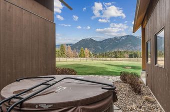 An outdoor hot tub with a brown cover is nestled between two modern rustic buildings, offering stunning views of snow-capped mountains and green fields under a blue sky.