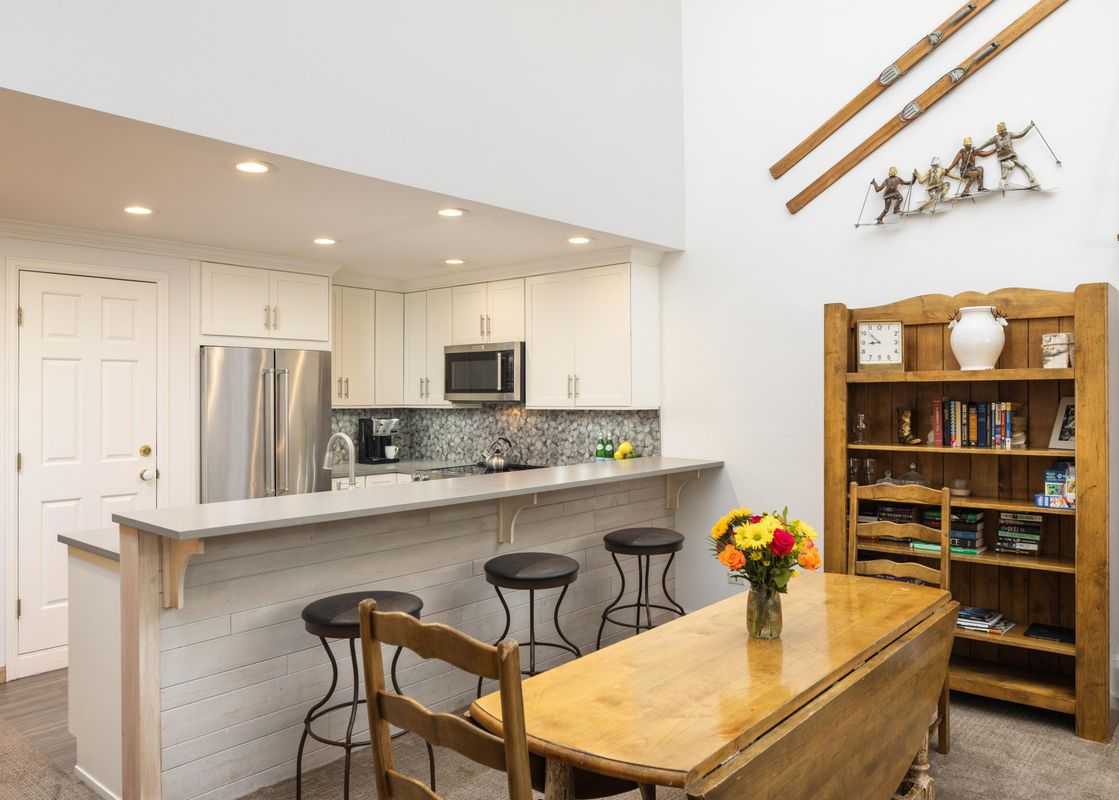 A modern kitchen with white cabinets, stainless steel appliances, a breakfast bar with three stools, and an adjacent dining table with a floral centerpiece.