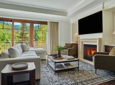 The spacious living area of the Montage Deer Valley One Bedroom Residence features a large sectional sofa, a coffee table, and a fireplace, with a window offering a lush green exterior view.
