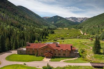 An aerial view of a large rustic log lodge nestled in a green mountain valley with a pond and winding road.