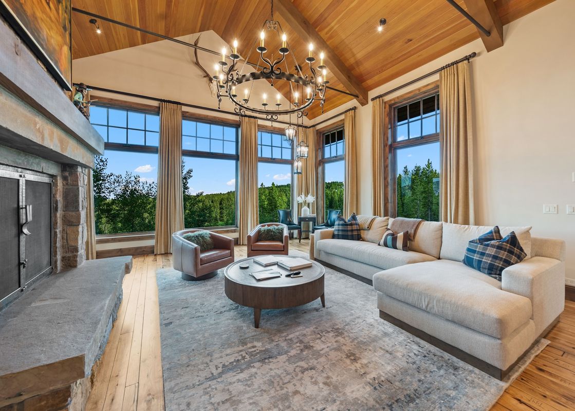 A spacious and elegantly furnished great room with large windows overlooking a forest, featuring a comfortable sectional sofa, armchairs, a coffee table, and a stone fireplace.