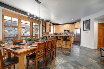 The image showcases the dining area and kitchen at Town Pointe A105, featuring a large wooden dining table, a modern kitchen with stainless steel appliances, and large windows offering scenic views.