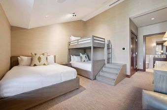 A bunk room with light wood furniture features a full bed and a bunk bed with white bedding and decorative pillows, alongside a nightstand and a partially visible door leading to a bathroom.
