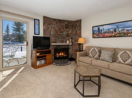 The living room of Meadow Ridge Court 9 Unit 4 features a stone fireplace, comfortable seating, and a view of the snowy outdoors.