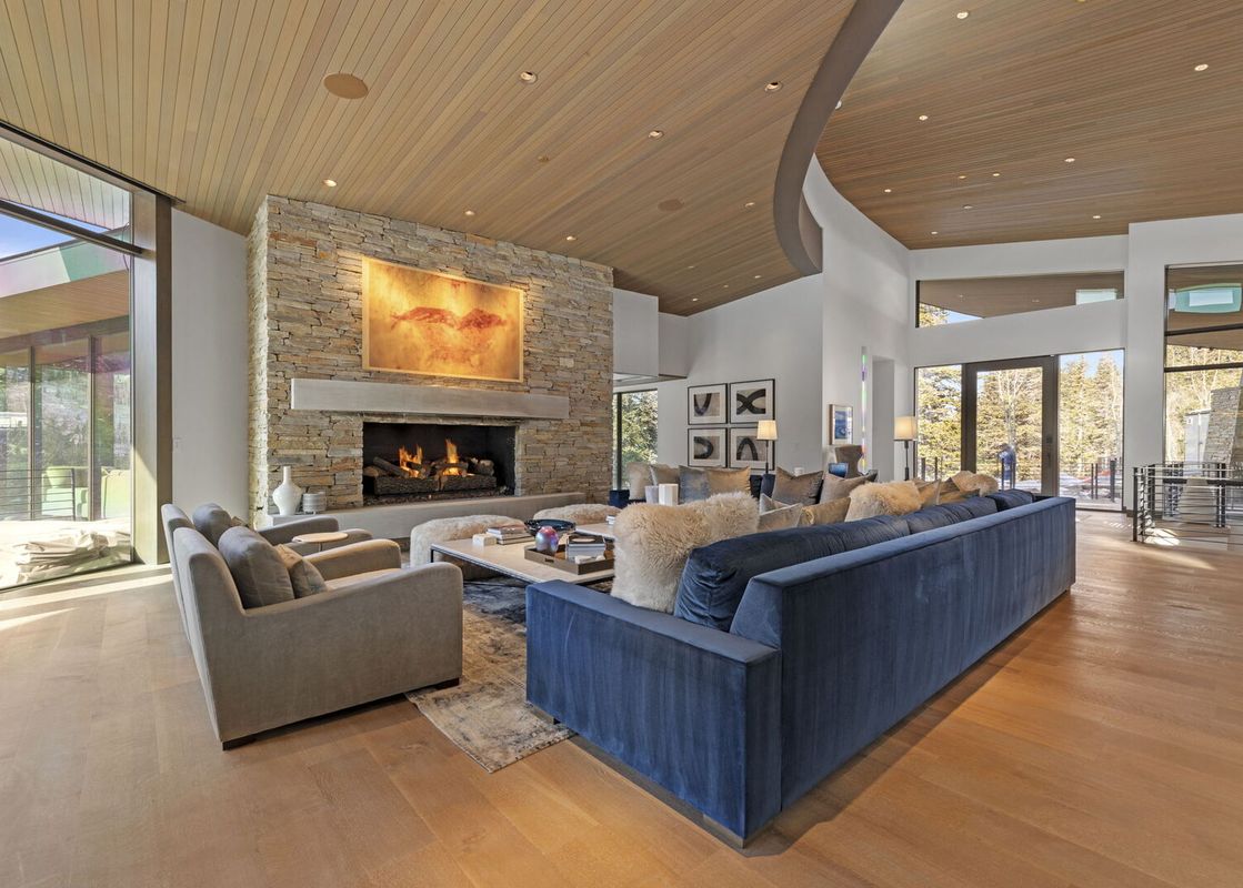 The spacious living area features a grand stone fireplace with a large painting above it, a deep blue velvet sofa, and light-colored armchairs arranged around a coffee table, all under a wood-paneled ceiling.