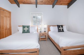 Two twin beds with white linens and green and black throw pillows are in a bedroom with wooden floors and a vaulted wooden ceiling.