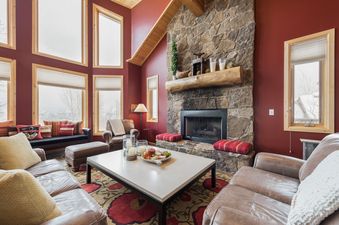 A luxurious and spacious living room at Solamere Home, featuring a large stone fireplace, comfortable leather seating, and expansive windows showcasing a winter landscape.