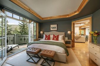 The master bedroom features a king bed with red and white bedding, two ottomans at the foot, and a private balcony overlooking a forest, with natural wood trim and dark gray walls.