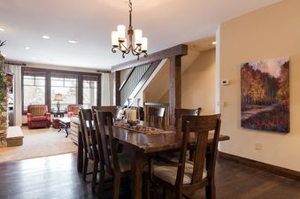 The dining area with a long wooden table and chairs is visible in the foreground, with a cozy living room featuring red armchairs and large windows in the background.