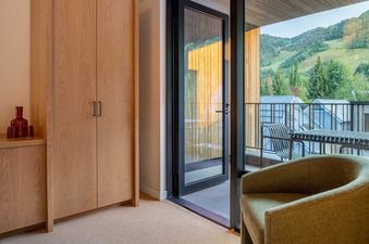 A modern room with light wood furniture, a cozy green chair, and a black-framed glass door opening to a balcony with outdoor furniture and a view of green mountains and houses.