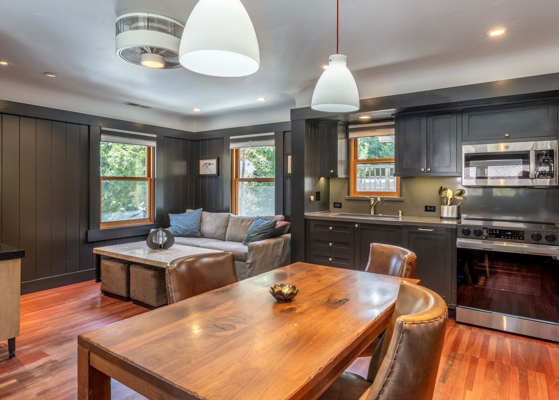 A modern open-concept living room and kitchen area with dark cabinetry, stainless steel appliances, a wooden dining table, and a comfortable sofa.