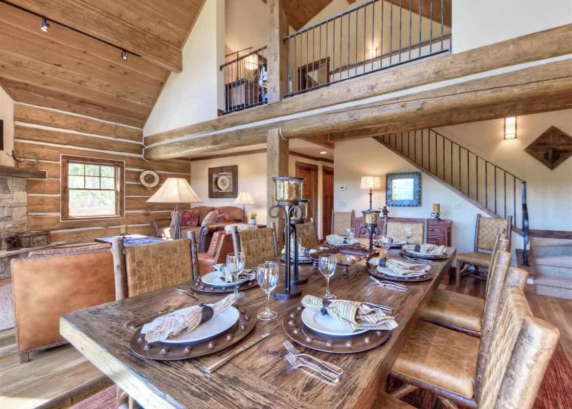 A rustic dining area with a long wooden table set for a meal, surrounded by leather chairs, and a living area in the background.