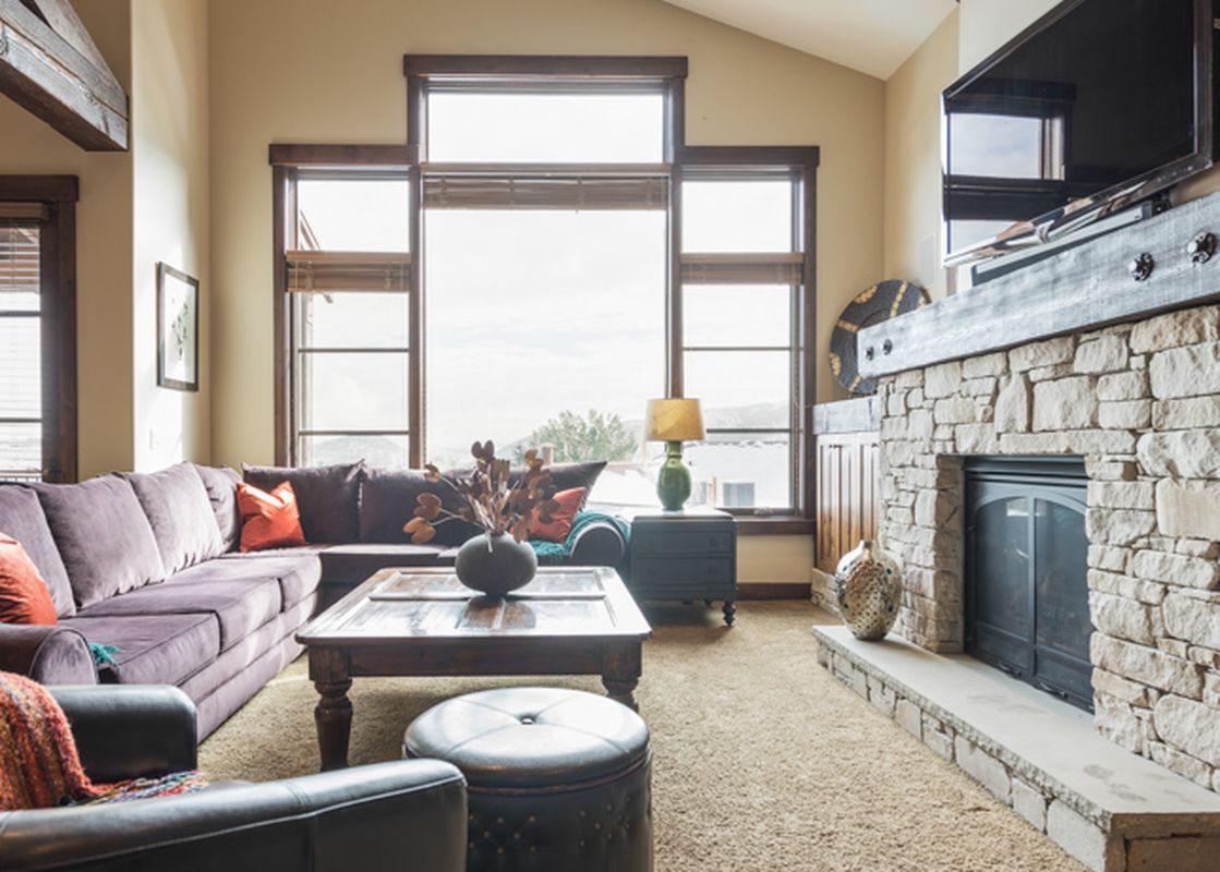An inviting living room at Silver Star 3302 featuring a large L-shaped purple sofa, a rustic coffee table, a stone fireplace with a TV, and large windows with mountain views.