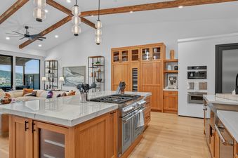 A modern kitchen with wooden cabinetry and a marble-topped island with a range, flowing into a living area with a view of hills through large glass doors.