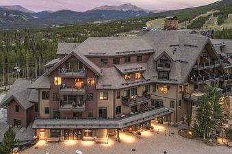 An aerial view of the Crystal Peak Lodge 7407 at dusk, showcasing its grand architecture, illuminated windows, surrounding pine forests, and distant mountains under a soft twilight sky.