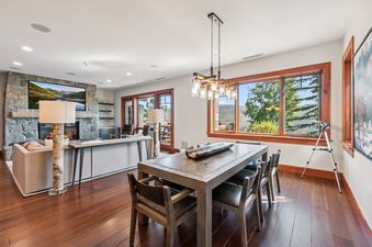 A dining room with a large wooden table and chairs, illuminated by a modern chandelier, and an adjacent living area with a stone fireplace and a large TV.