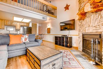 The spacious living room at Powder Pointe 303B, featuring a gray sectional, wooden coffee table, stone fireplace, flat-screen TV, and an open kitchenette with wooden cabinets.