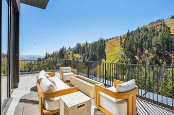 A spacious private balcony at Founders Place 1704, furnished with modern outdoor seating and a fire pit, overlooks a stunning mountain landscape with green and yellow trees under a clear blue sky.