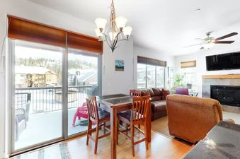 The image shows the interior of a luxurious vacation rental unit, featuring a dining area, living room, and large windows that offer a view of the snowy mountains outside.
