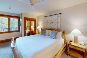 A bedroom with a king-sized bed, a large window with a view of the trees outside, and a wooden nightstand with a lamp.
